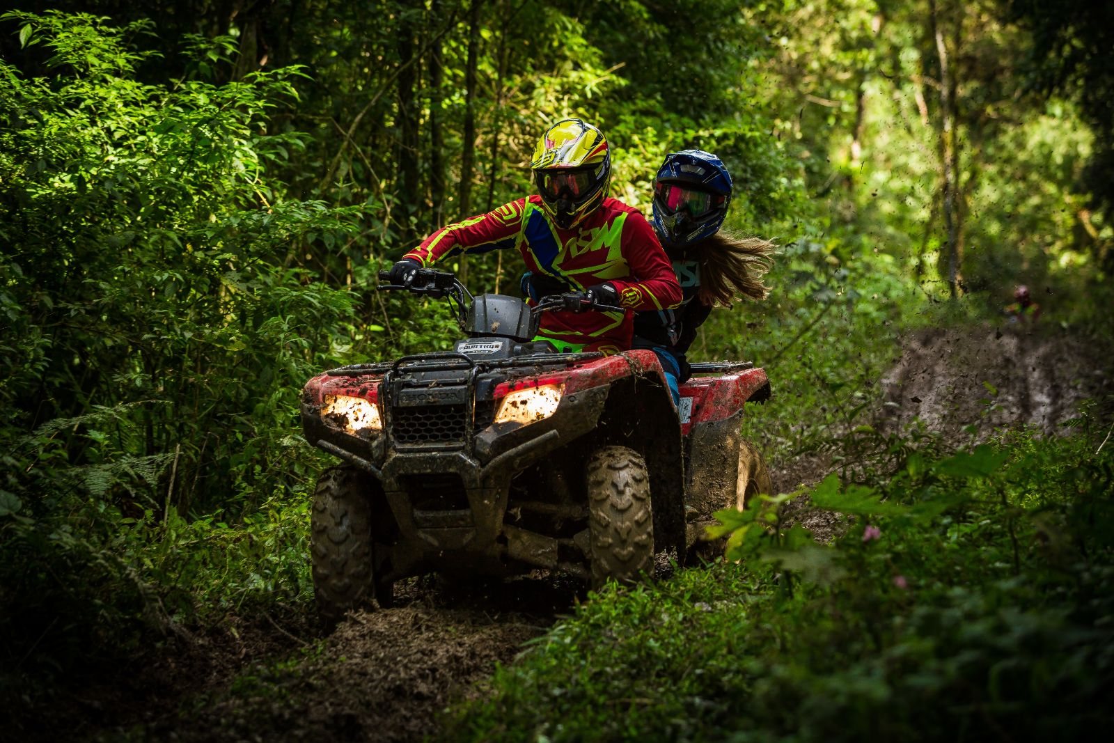 A couple on an ATV driving through the dense forest trees in the Slave Lake Region of Northern Alberta, with helmets and protective riding gear and ATV headlights towards the camera
