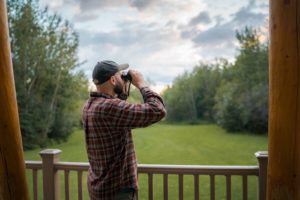 a man in plaid and a baseball cap looking through binoculars, standing on a wood-railing deck, looking out over a grassy field and trees