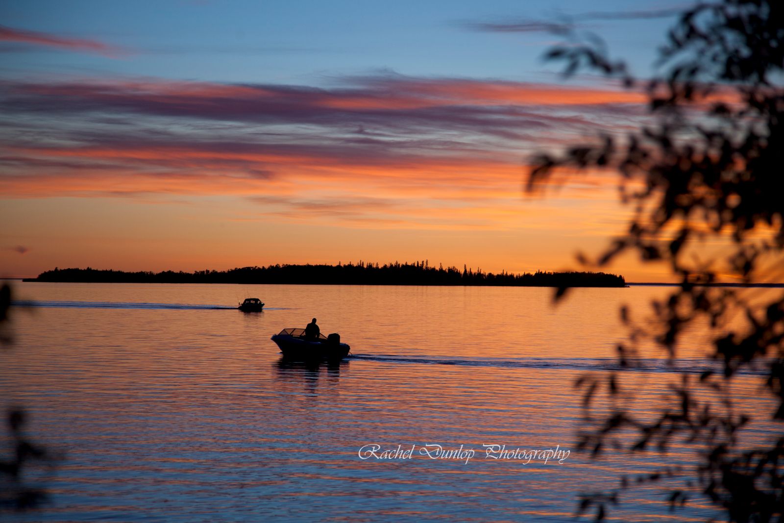 Two boats at sunset in the Slave Lake Region of Northern Alberta, shown as dark silhouettes against an orange, pink and purple sunset, with an island silhouette in the distance