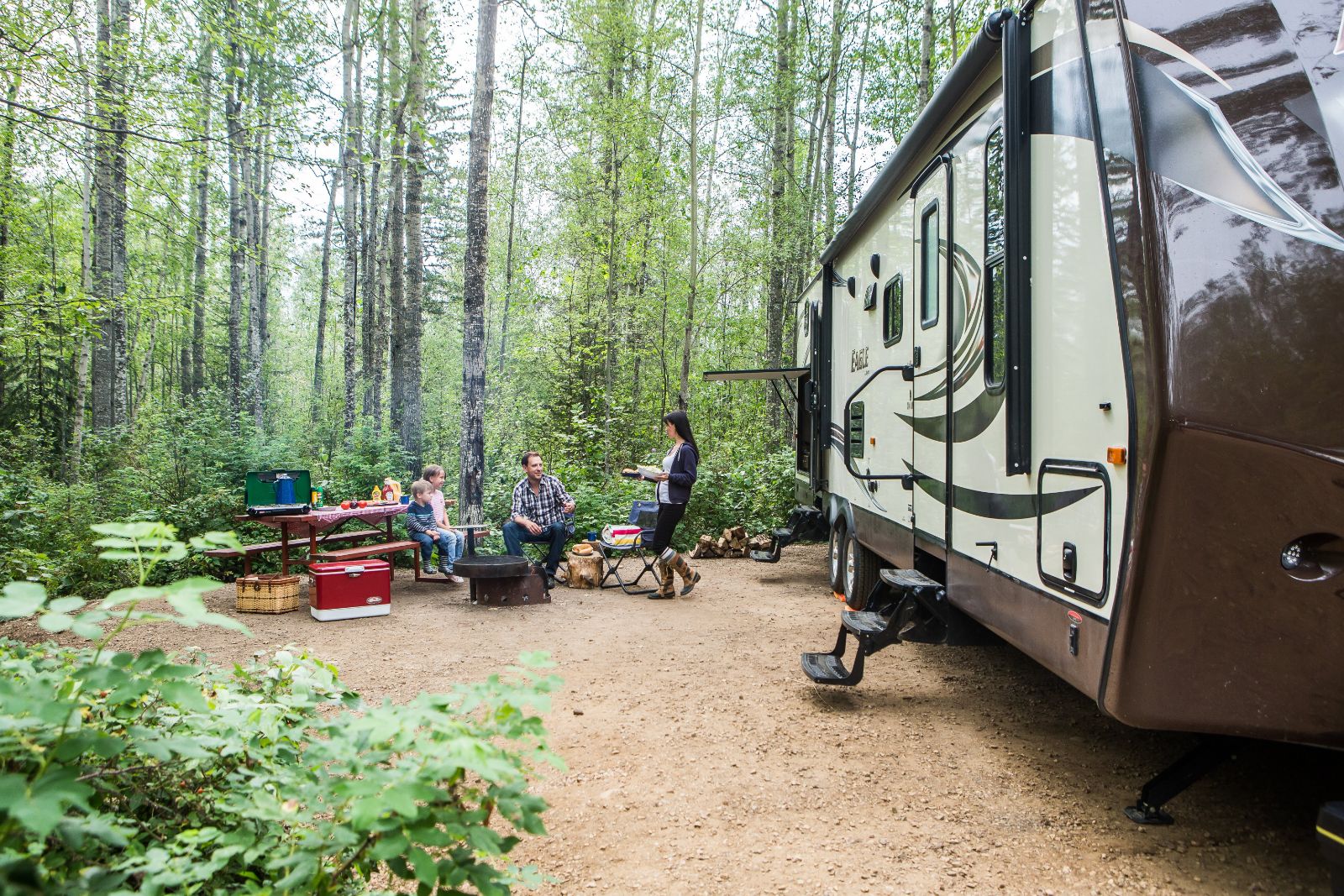 An RV camper at a campground in the Slave Lake Region of Northern Alberta, with a family sitting in camping chairs around a campfire between the trees
