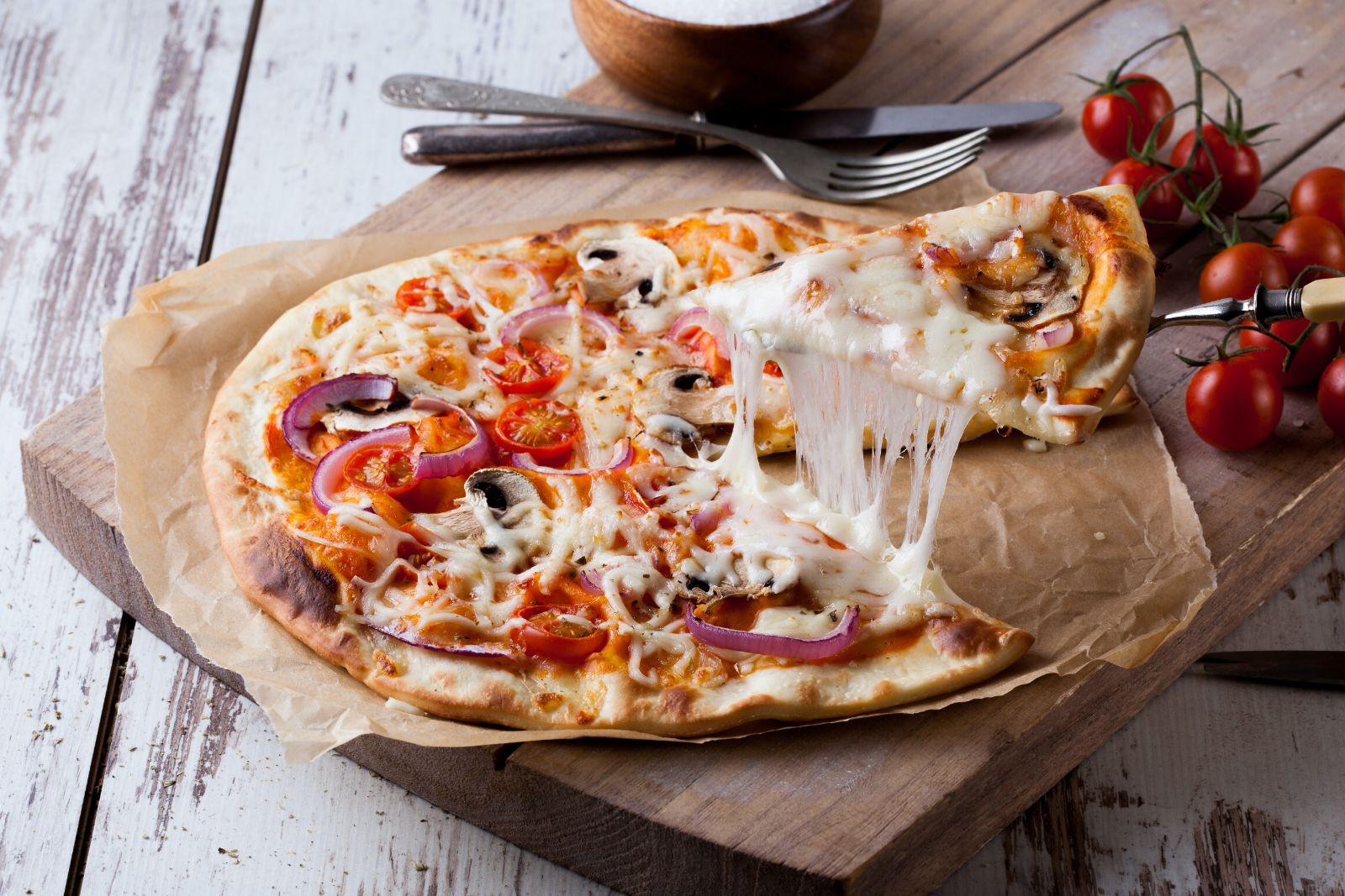A cheesy slice of pizza being lifted from the whole pizza with mushrooms, onions, tomatoes and sauce on a thin crust