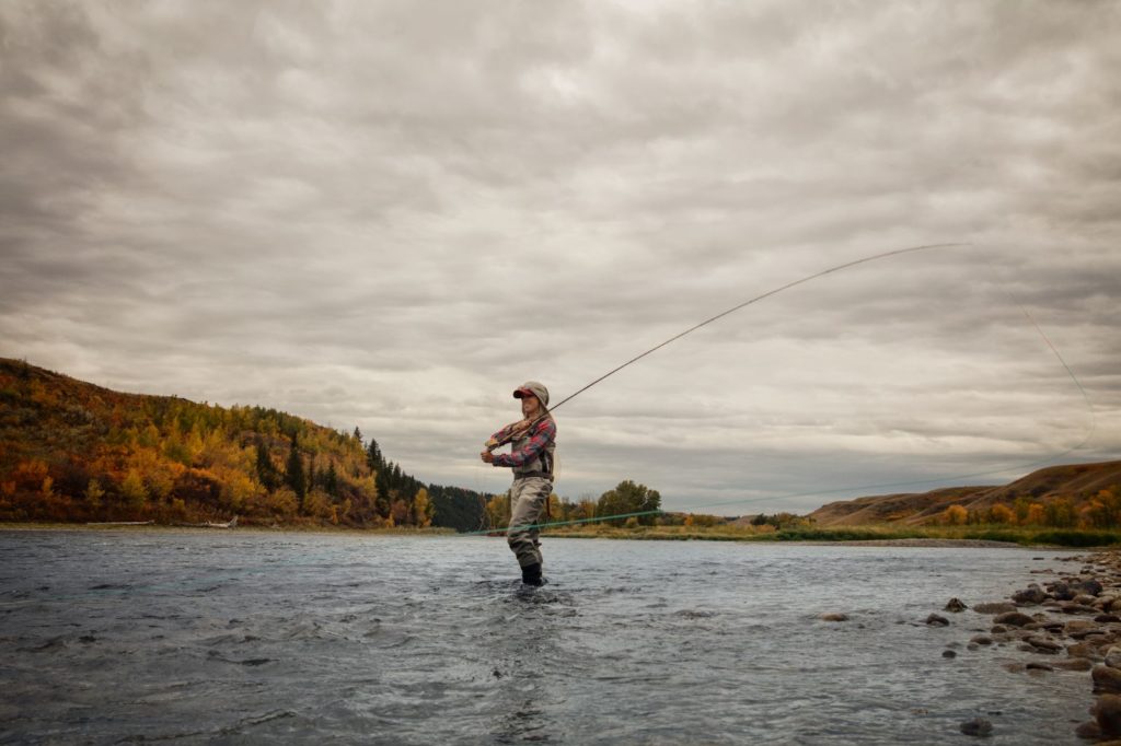 A person fly fishing at Lesser Slave Lake, waded into the water in their fishing gear, with the lake and mountains and cloudy sky in the background
