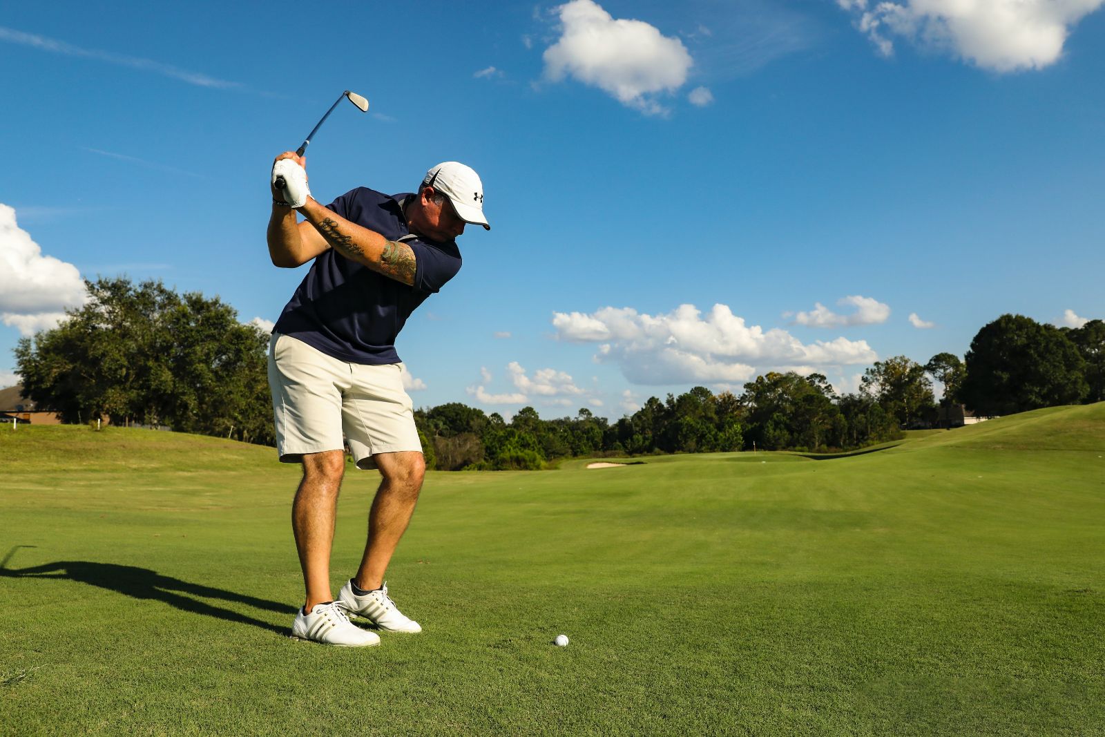 man in a golf shirt, shorts and a white hat, swinging a golf club at a ball at The Gilwood Golf Club; greens, trees and blue skies with white clouds in the distance