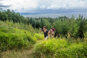 Two young people hiking in the Slave Lake Region, with green bushes and trees surrounding them on a walking trail, light clouds and mountains in the distance