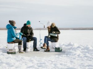 three people dressed in winter gear seated around an ice fishing hole with snow surrounding them at Lesser Slave Lake