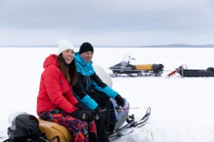 a couple sitting on a snowmobile, dressed in warm winter gear, looking back at the camera smiling, with another snowmobile in the background