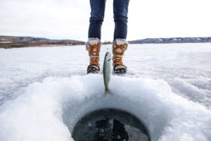 A closeup of an ice fishing hole with a small fish being pulled out of the hole and a fisherman's winter boots in the background on an iced-over lake